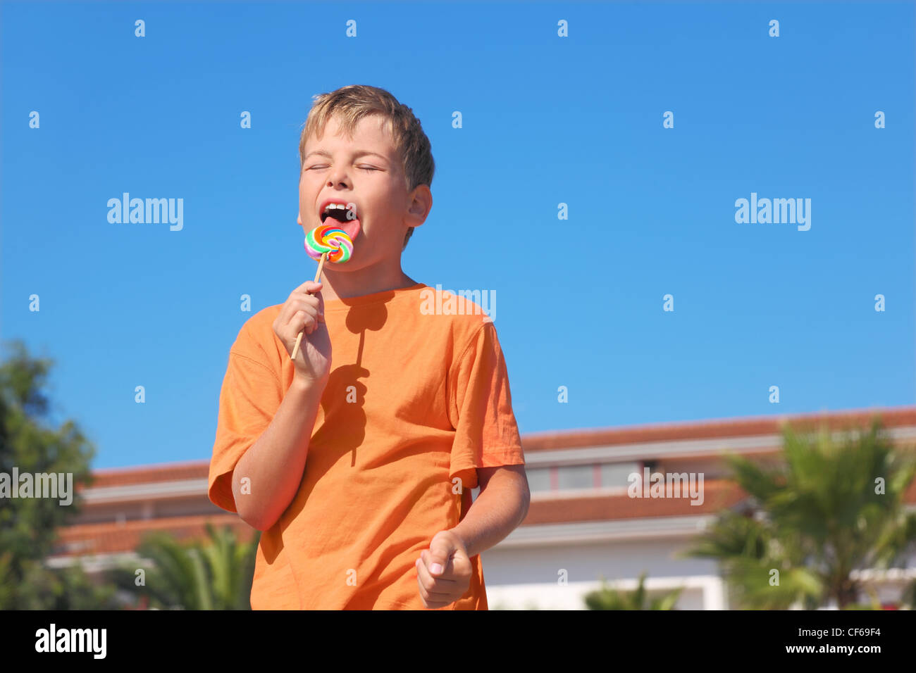 little boy in orange shirt licking multicolored lollipop, closed eyes, palms and building Stock Photo
