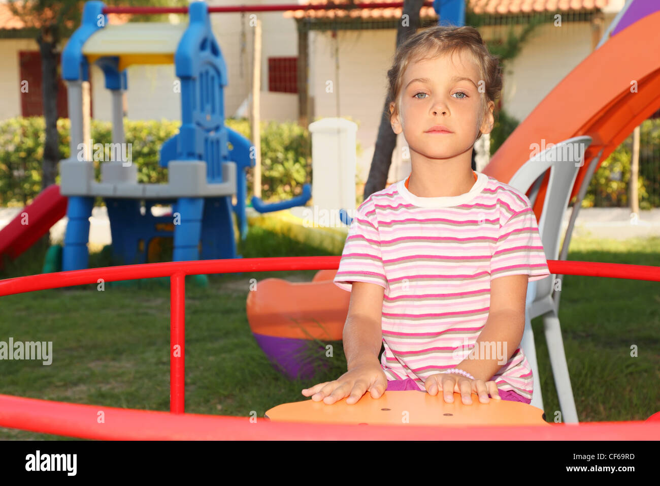 serious little girl in pink shirt sitting on merry-go-round on playground and looking at camera, building Stock Photo