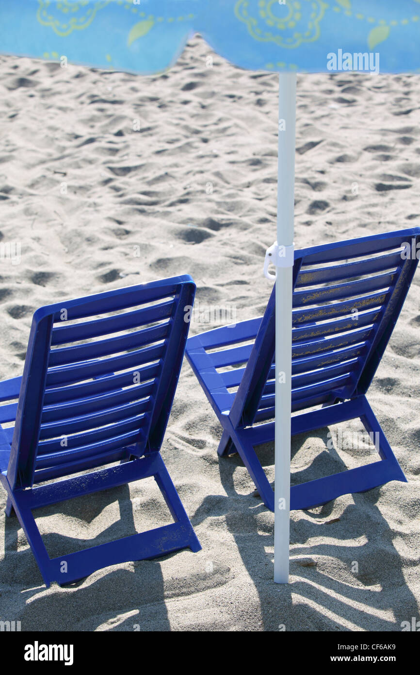 Two dark blue plastic chairs stand on  beach under  umbrella Stock Photo