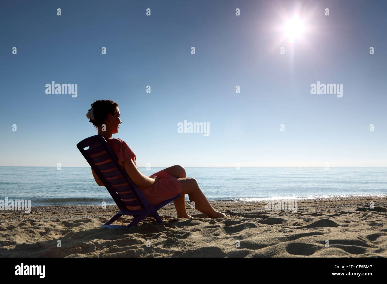 Woman sits on  dark blue plastic chair sideways and becomes tanned in  morning on  beach Stock Photo