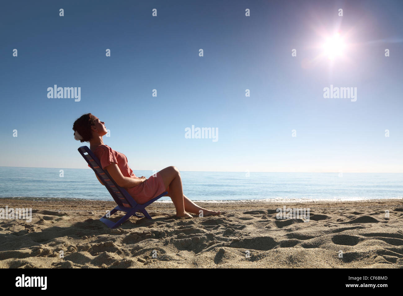 Woman sits on  plastic chair on  sea-shore by  person to  sun and becomes tanned, heaving up  head Stock Photo