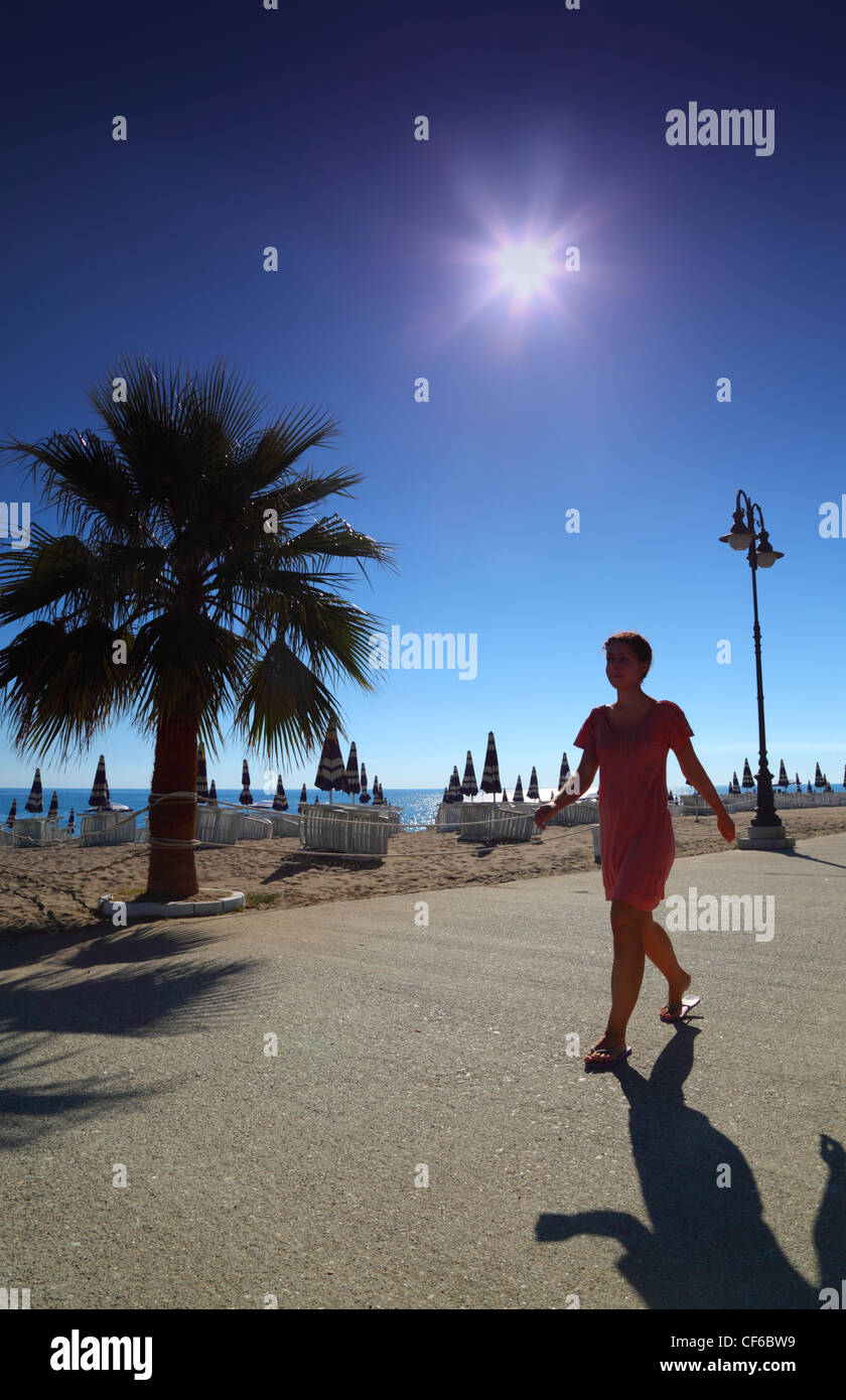 Girl goes on path with palms on empty sandy beach with folded umbrellas and sunbeds, burning sun and cloudless sky Stock Photo
