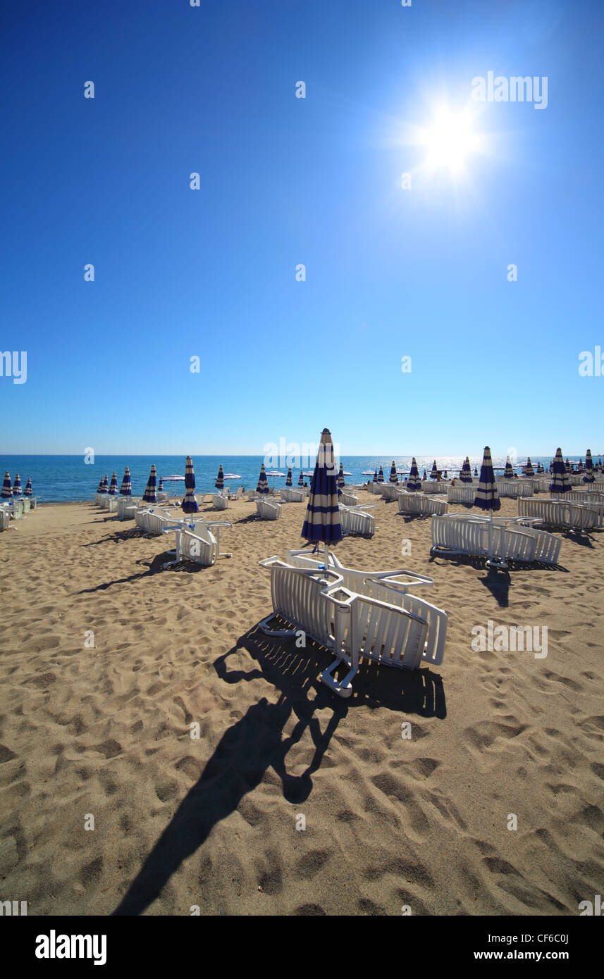 Empty sandy beach with folded and open umbrellas and sunbeds, burning sun and cloudless sky Stock Photo