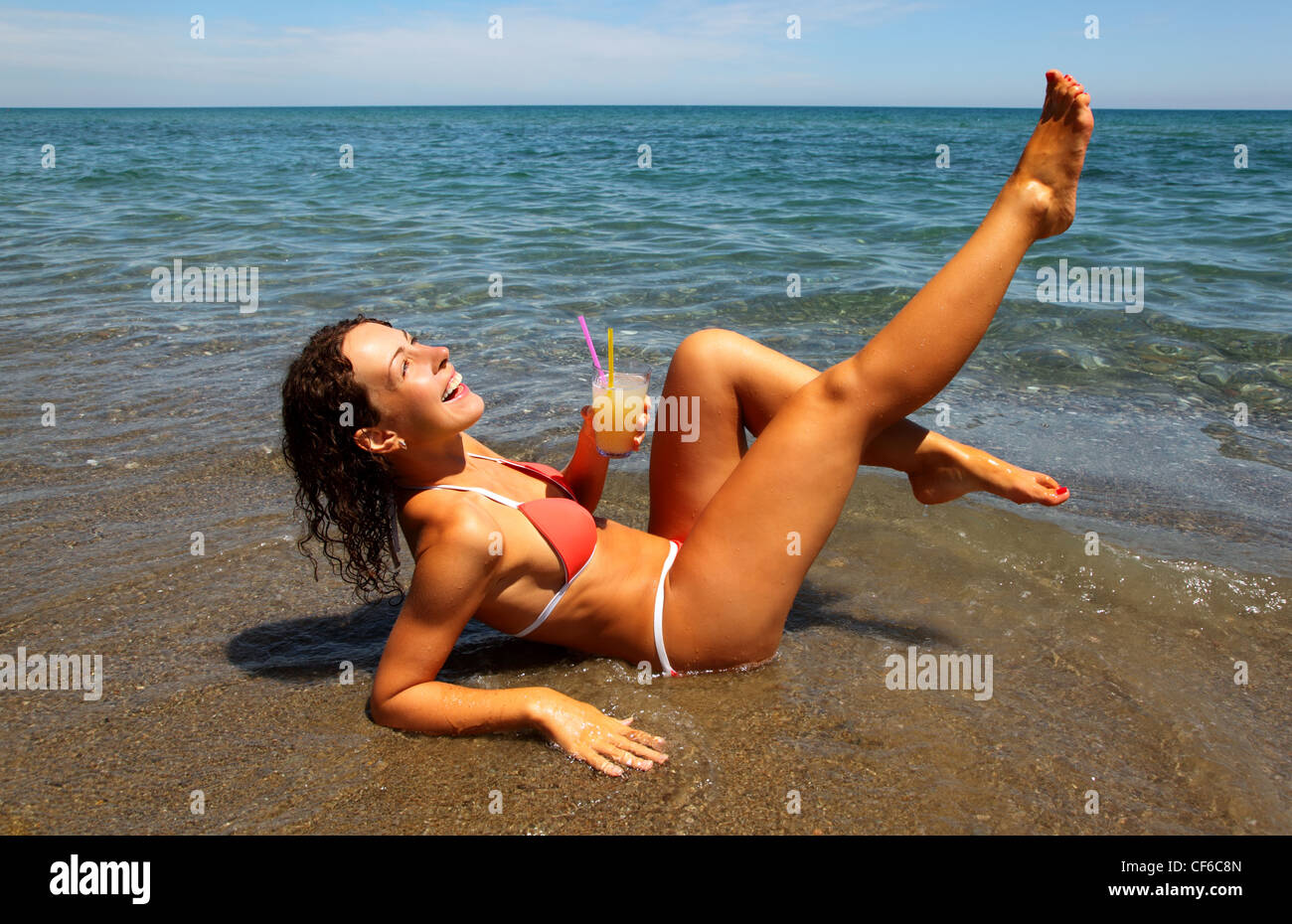Girl in red swimsuit lie in sea. She holds a glass of juice with two tubes. Stock Photo