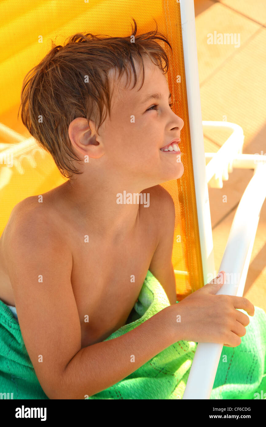Cheerful little boy sits in yellow deck chair and wrap oneself up by green coverlet Stock Photo