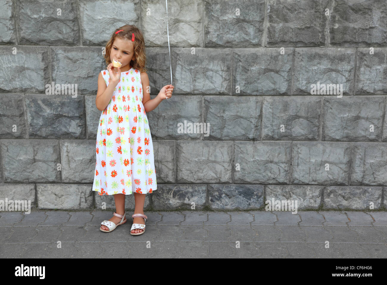 Little girl in  white sarafan eats an ice-cream and holds an air marble near  stone wall Stock Photo