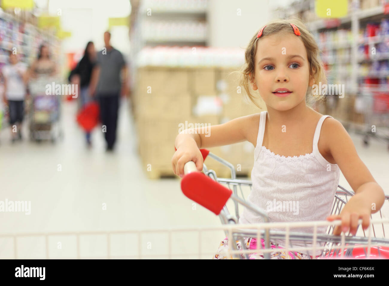 little girl sitting in store cart and looking at side, shelves with commodity Stock Photo