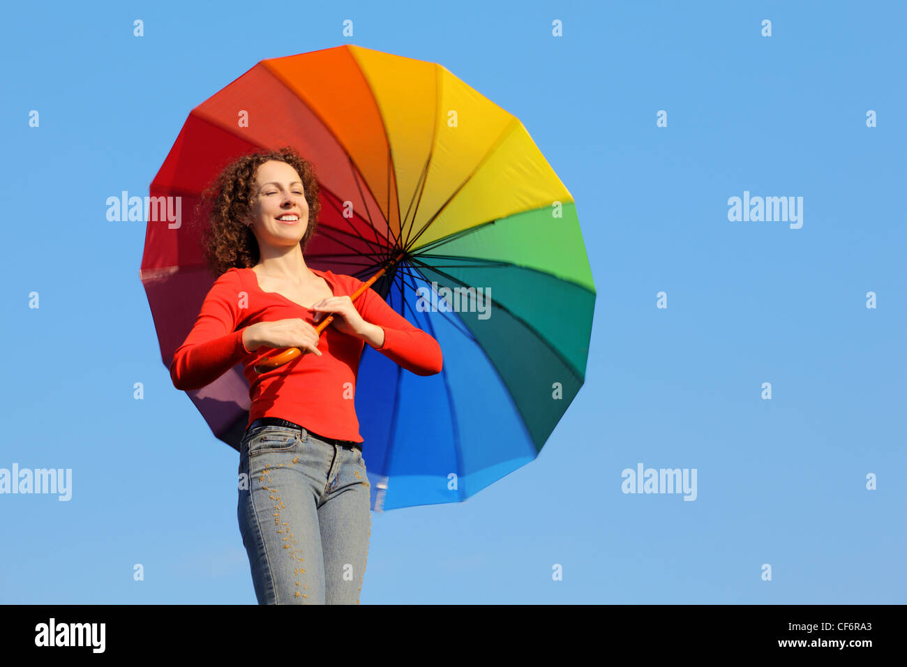 Girl with eyes closed because of glare of sun stands against blue sky and holding colored umbrella Stock Photo
