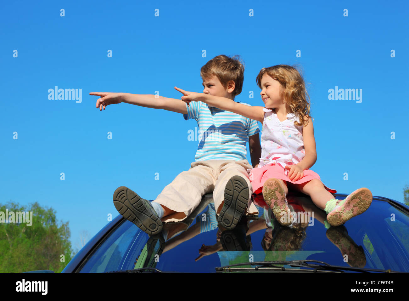 little boy and girl sitting on car roof on blue sky, looking to left ...