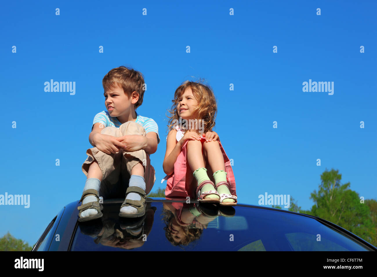 little boy and girl sitting on car roof on blue sky, hands on legs ...
