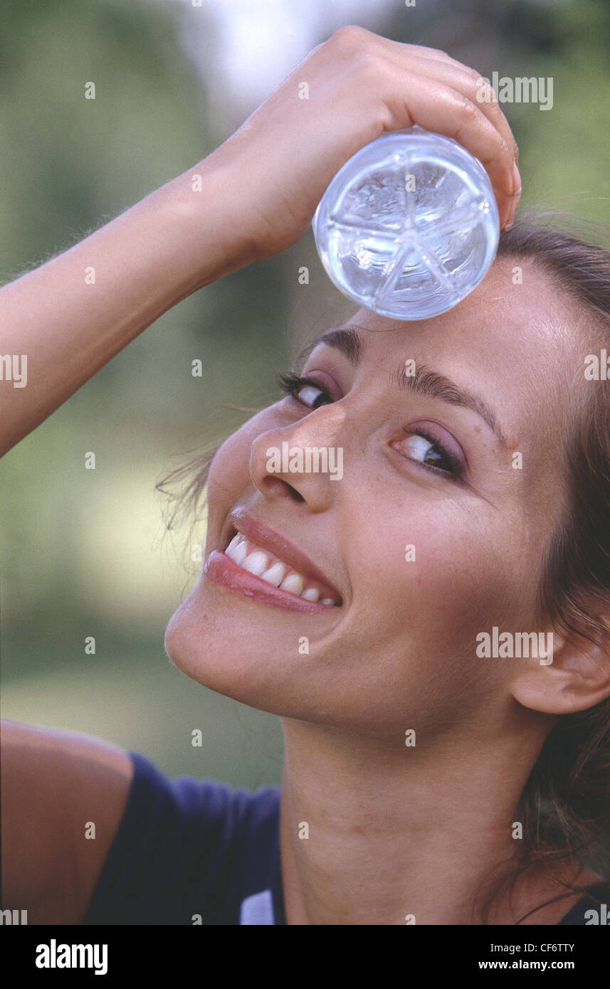 Semi profile of female long brunette hair in ponytail wearing subtle make up and blue top, holding bottle of water to forehead Stock Photo