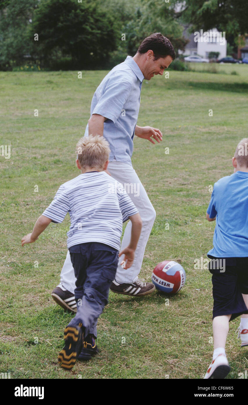 Male short brown hair playing football in park two male children blonde hair  running towards ball away from camera Stock Photo - Alamy