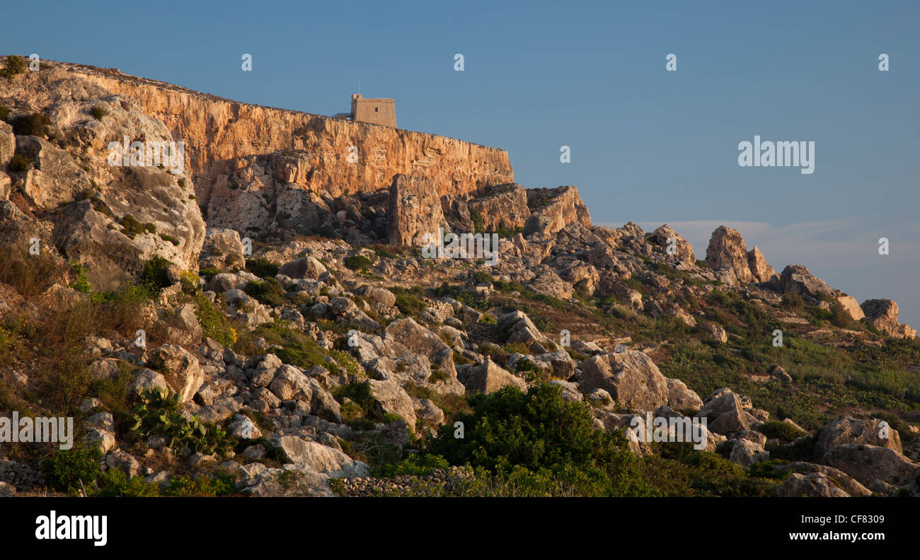 A medieval coastal watchtower stands out on top of a bluff looming over slopes ringed with boulders in Gozo's north coast. Stock Photo