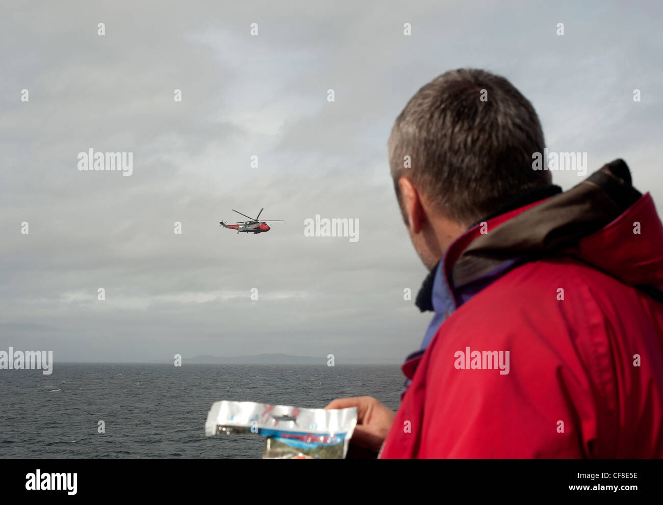 A man in red jacket watches an RAF Sea King rescue helicopter hover close by above the sea in Scotland.. Stock Photo