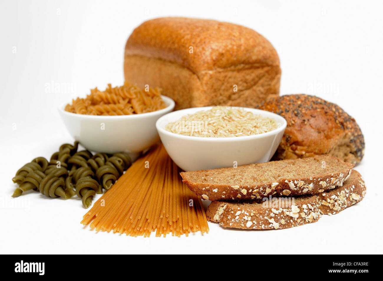 Two bowls filled long grain rice and dried fusilli pasta surrounded by loaves of bread, dried spaghetti and dried spinach Stock Photo