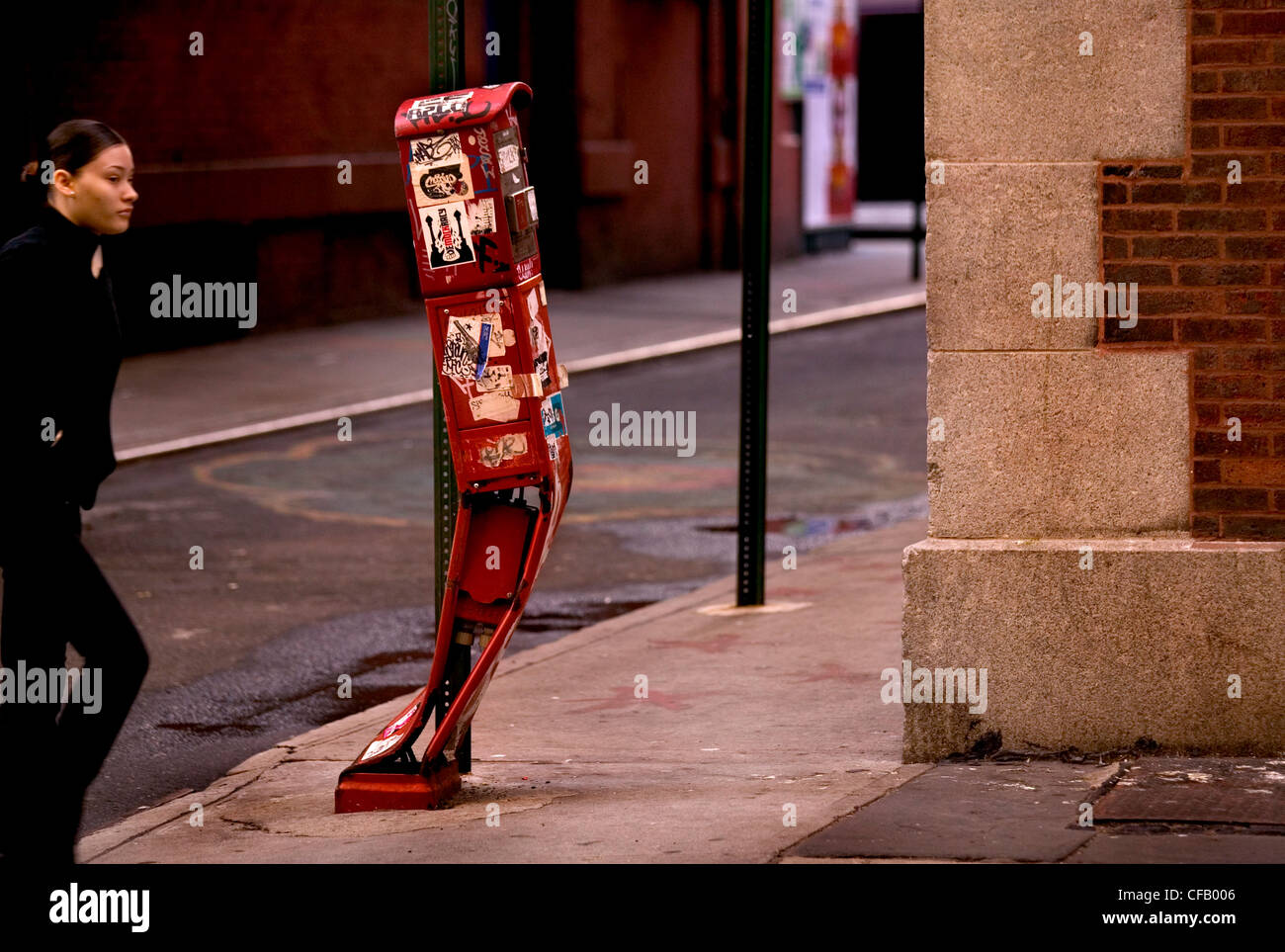 A woman walking past a bent red letter box which is covered in stickers and graffiti. Stock Photo