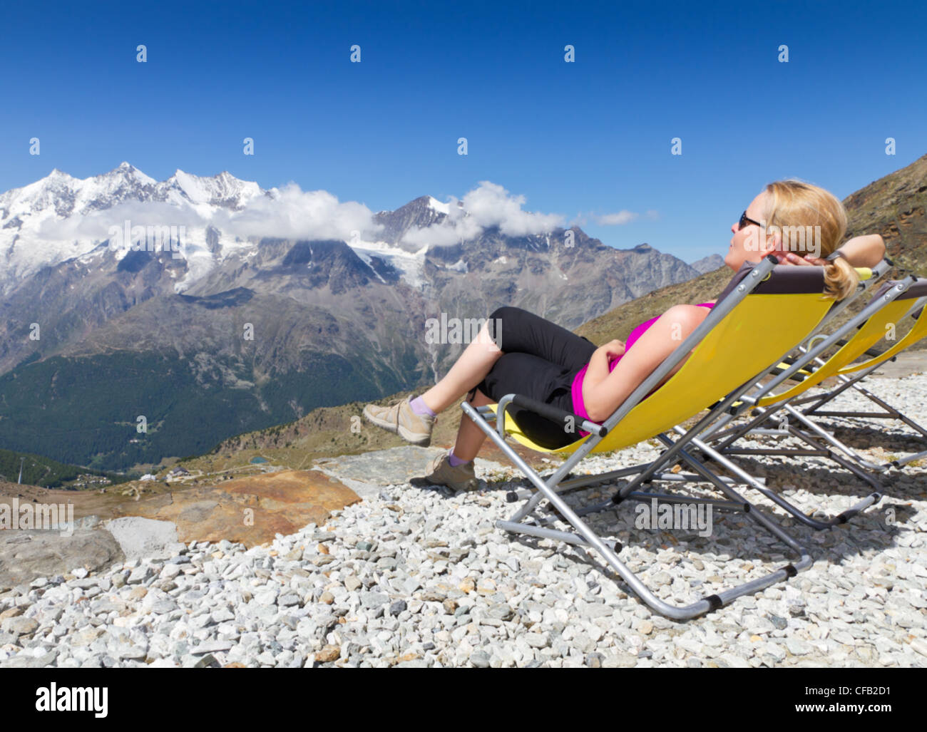 young woman enjoys the sun and relaxes on a canvas chair surrounded by alpine snow capped mountains Saas Fee, Switzerland Stock Photo