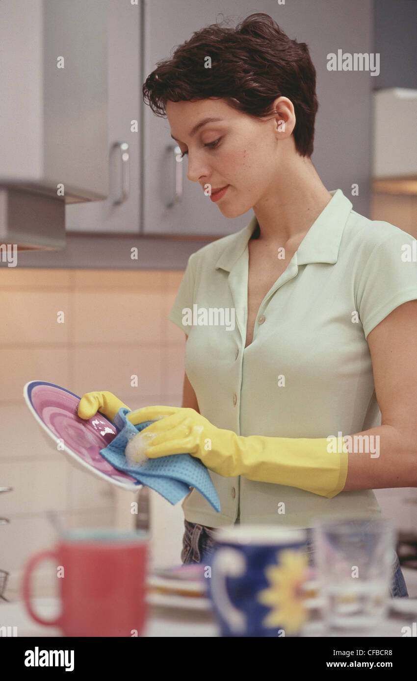 Semi profile of female short brunette hair wearing red lipstick pale green blouse blue jeans and yellow rubber gloves, washing Stock Photo