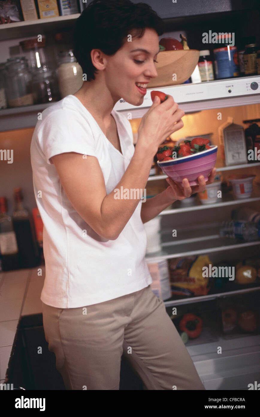 Semi profile of female short brunette hair wearing white t shirt and beige trousers, in kitchen next to open fridge eating Stock Photo