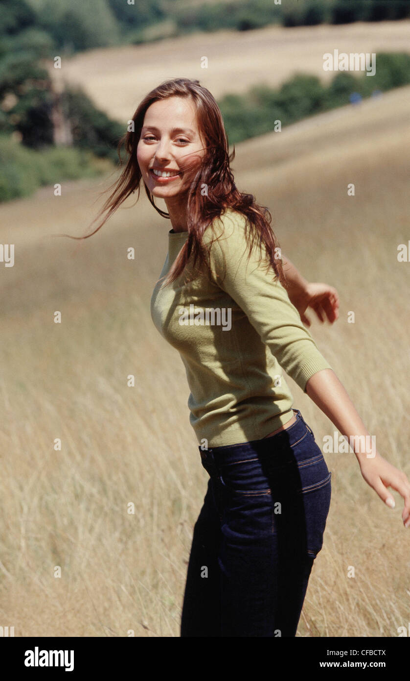 Semi profile of female long brunette hair wearing subtle make up green top and blue jeans standing in field arms outstretched Stock Photo