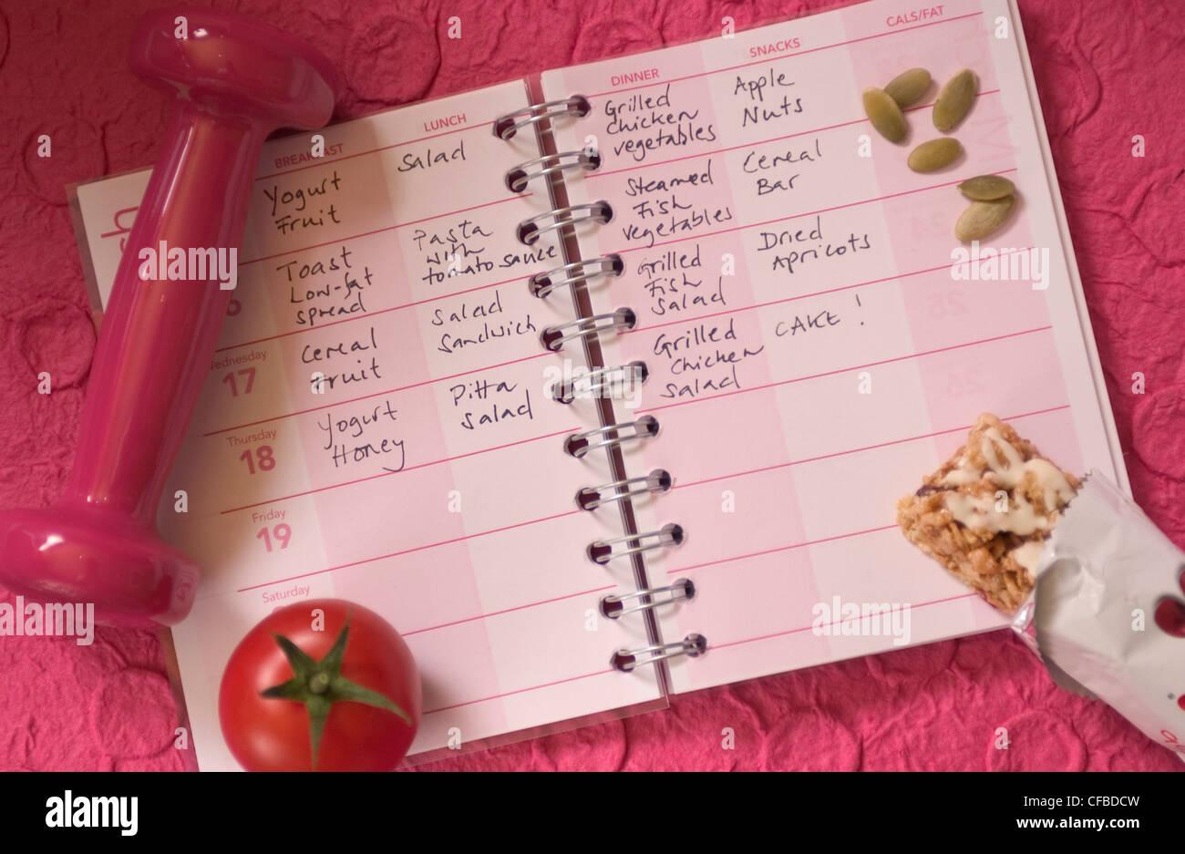Diet diary with pumpkin seeds, dumb bell, cereal bar and tomato on background of pink embossed paper Stock Photo