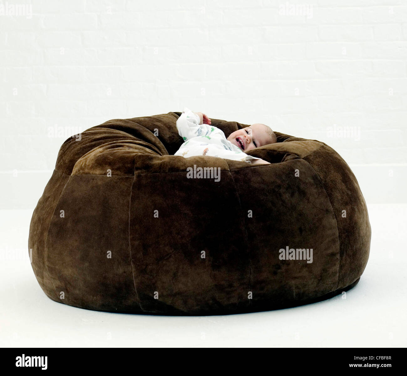 Male baby wearing white patterned babygro, lying on back on large brown suede beanbag looking to distance smiling, against Stock Photo