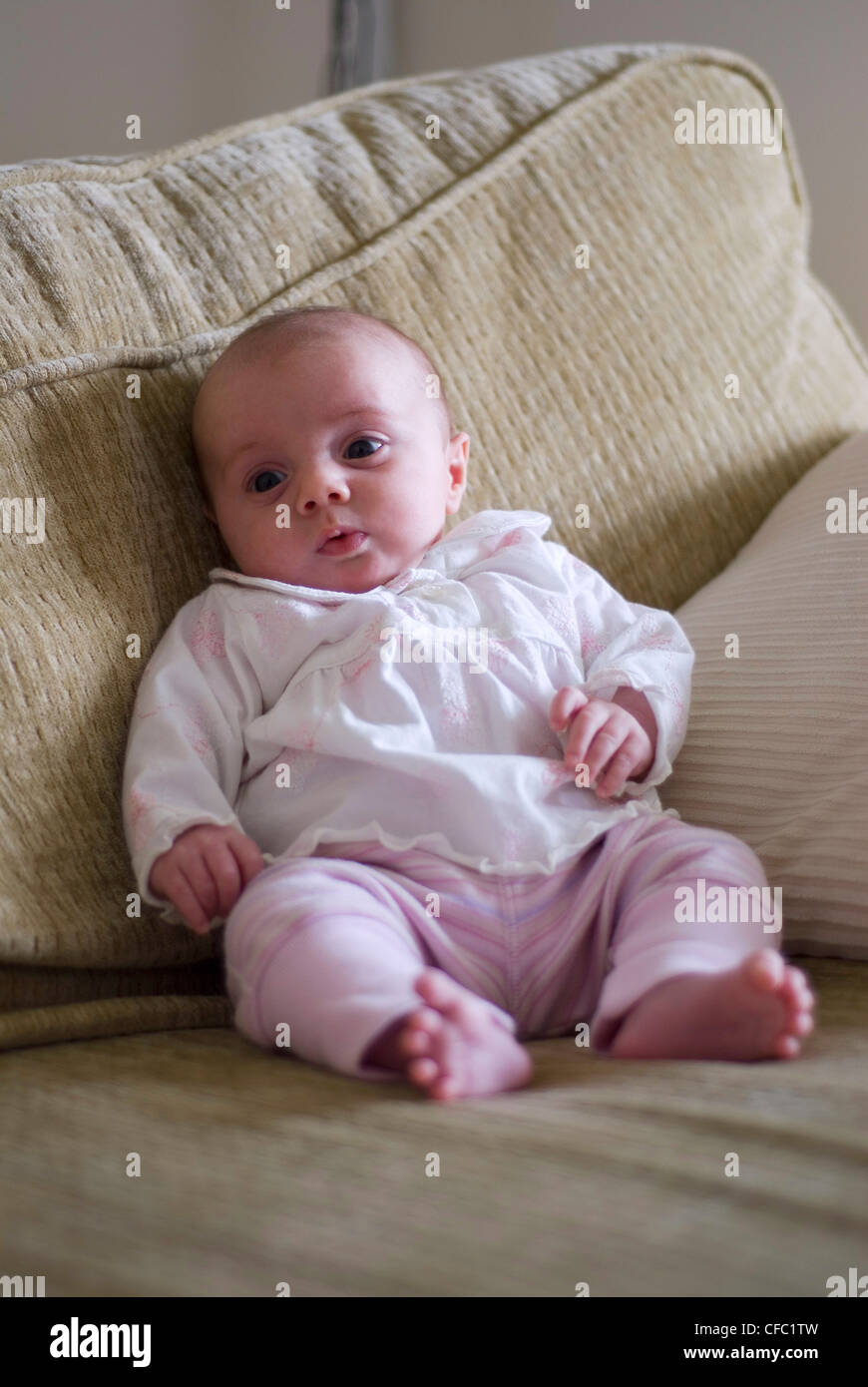 Female two month old baby propped up on a beige sofa, dressed in a