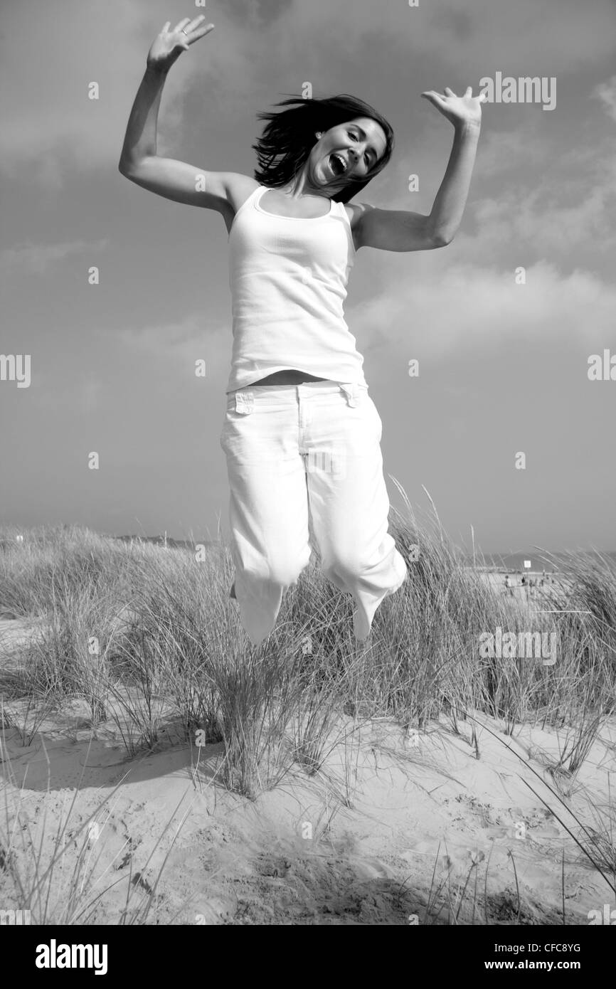 Female shoulder length brunette hair wearing a white vest and white trousers, jumping on the beach, legs bent in knees, arms Stock Photo