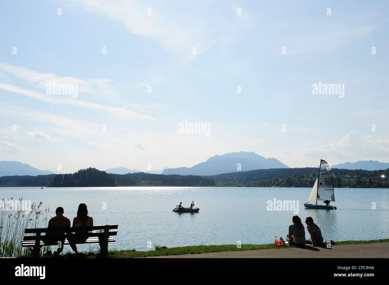 Bathers sitting at lakeside of lake Faaker See with rowing boat and sailing boat, lake Faaker See, Carinthia, Austria, Europe Stock Photo