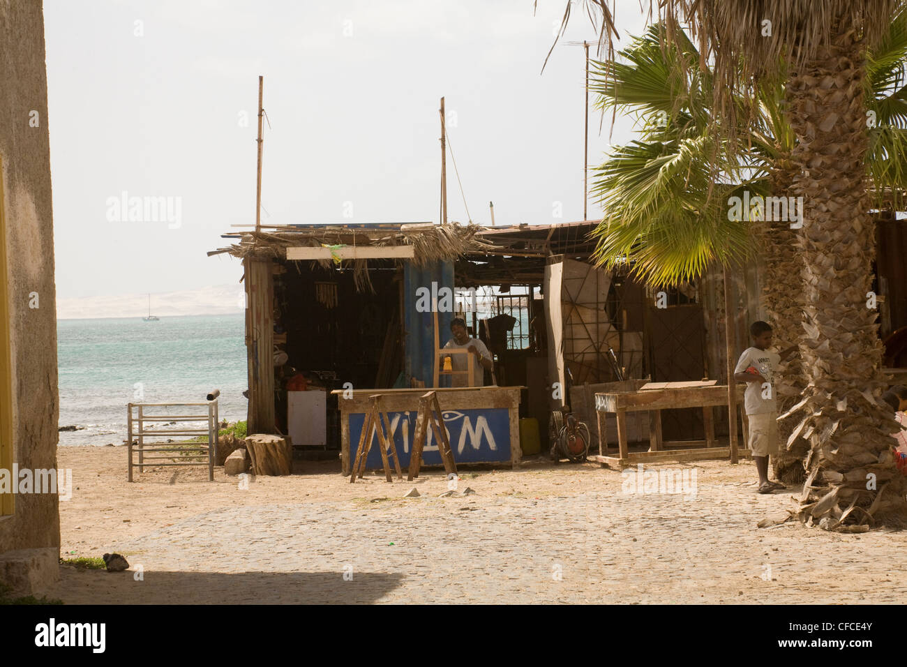 Sal Rei Boa Vista Cape VerdeLocal man building a chair in a water front carpenters' shack young boy playing around palm tree Stock Photo