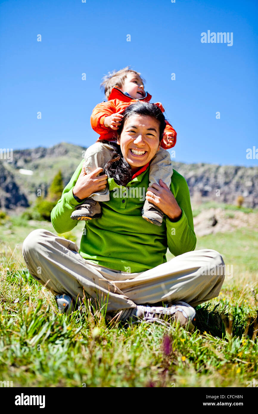 a woman smiles as she sits cross legged in an alpine meadow with wildflowers doing a Buddha pose. Her son, a 2 year old, crawls Stock Photo