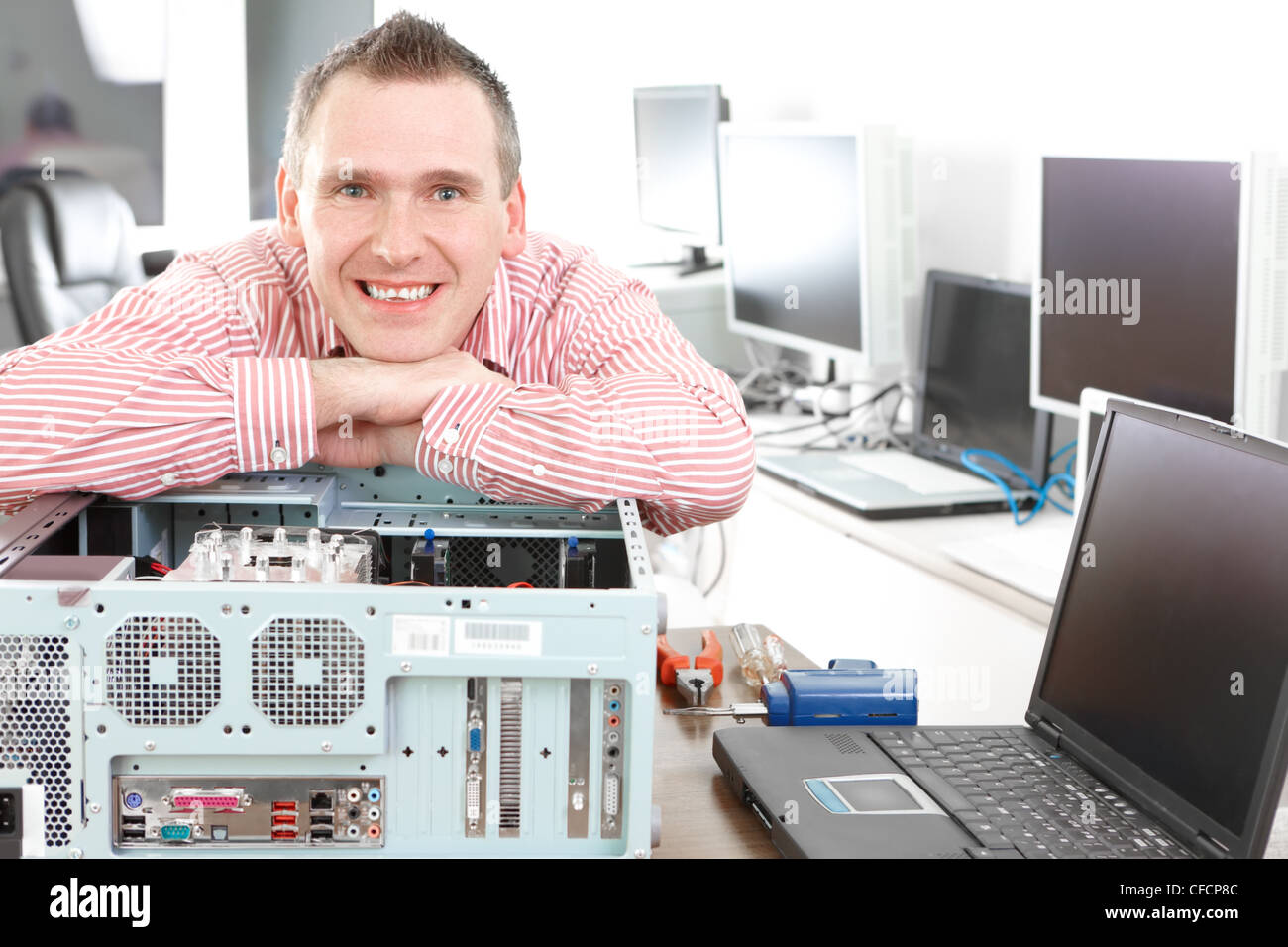 Repairman with computer, an owner of small business. Monitors and other laptops in the background waiting for service. Stock Photo