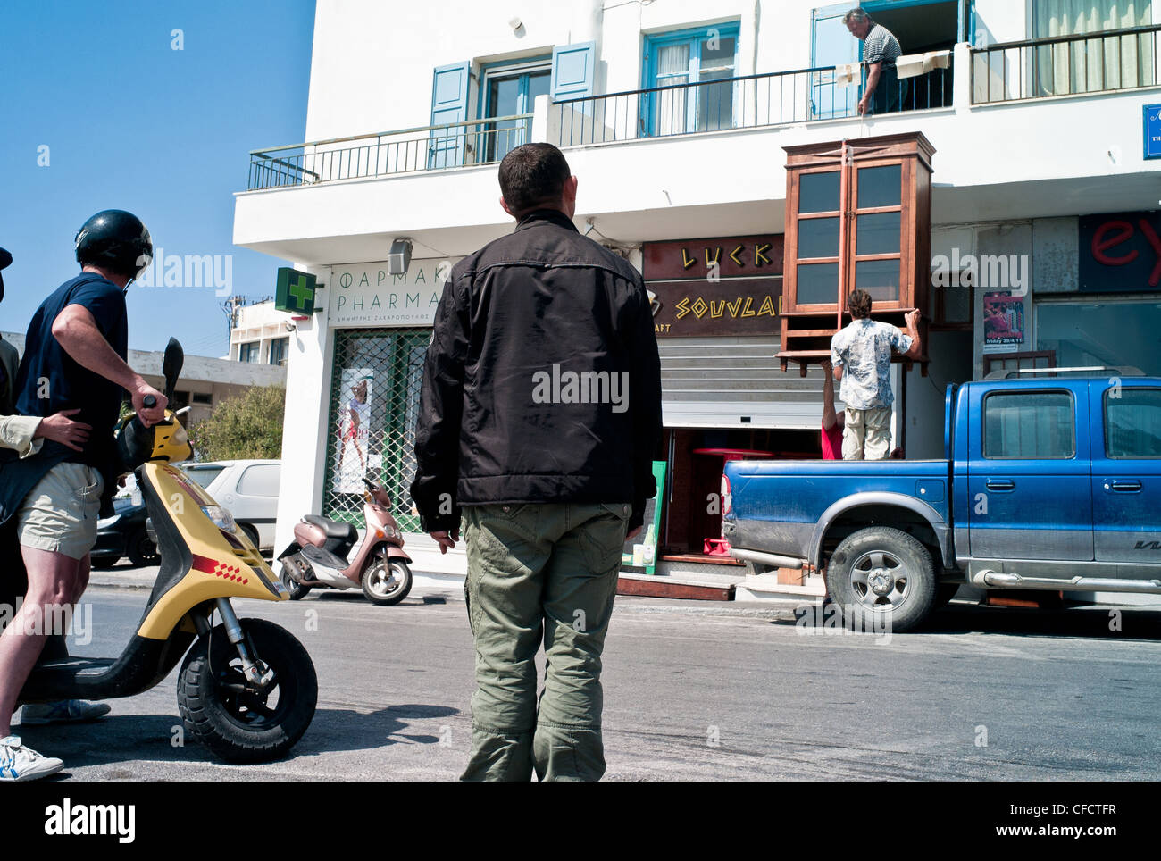 A man watches two men removing a piece of furniture from a balcony into a pick up truck on a Greek Island. Stock Photo