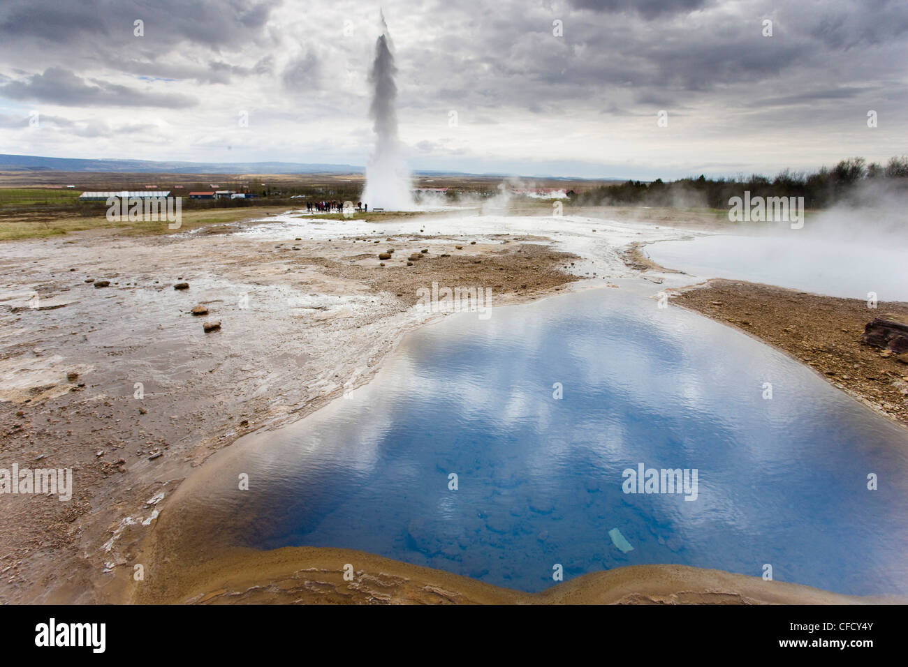 Blue water in geothermal pool with water spout from Strokkur Geysir visible in the distance, Geysir, near Reykjavik, Iceland Stock Photo