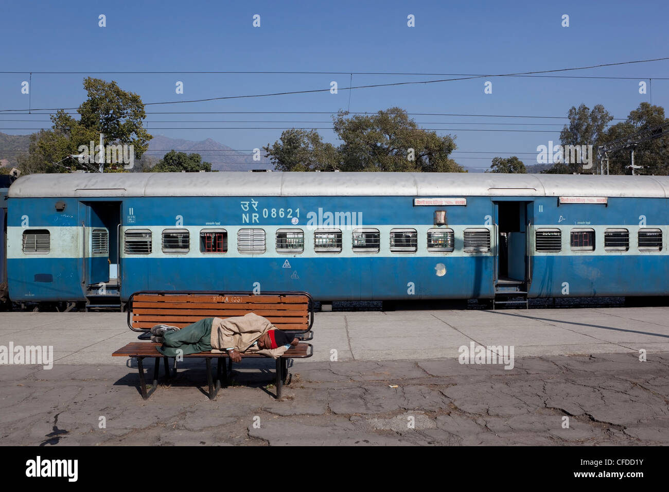 a man sleeps on a bench at Kalka railway station in the Indian state of Haryana, with blue railway carriages Stock Photo