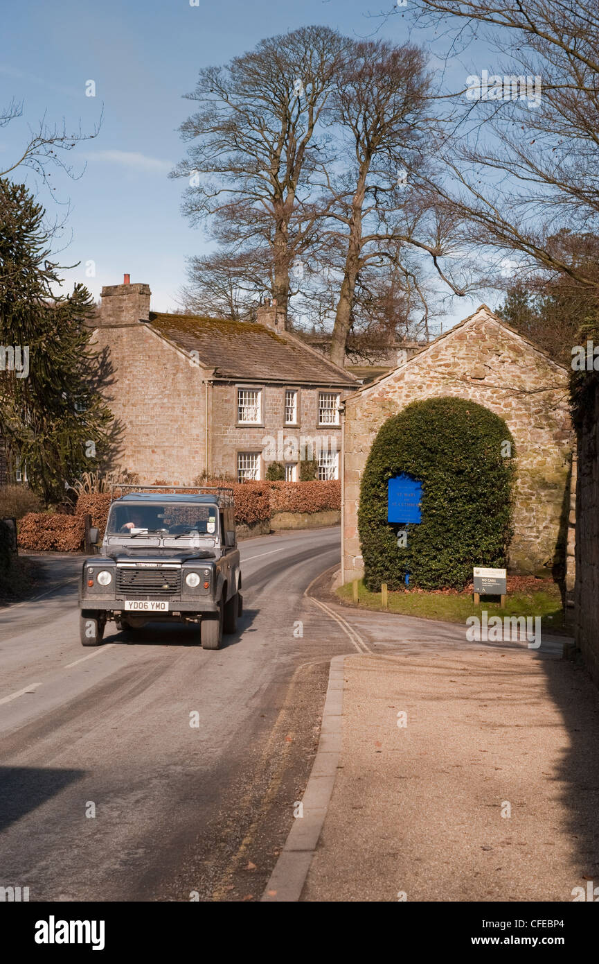 Front view of Land Rover Defender traveling or being driven on country road past quaint cottage - Bolton Abbey village, North Yorkshire, England, UK. Stock Photo