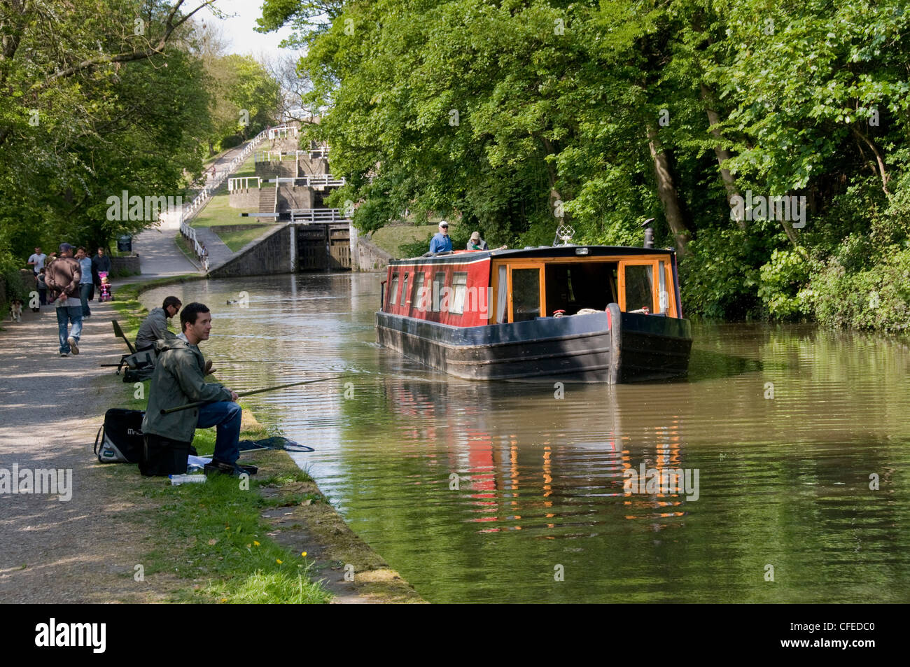 Narrowboat travels on scenic waterway (Five Rise Locks, Leeds Liverpool Canal - couple aboard) anglers & walkers - Bingley, W Yorkshire, England, UK. Stock Photo