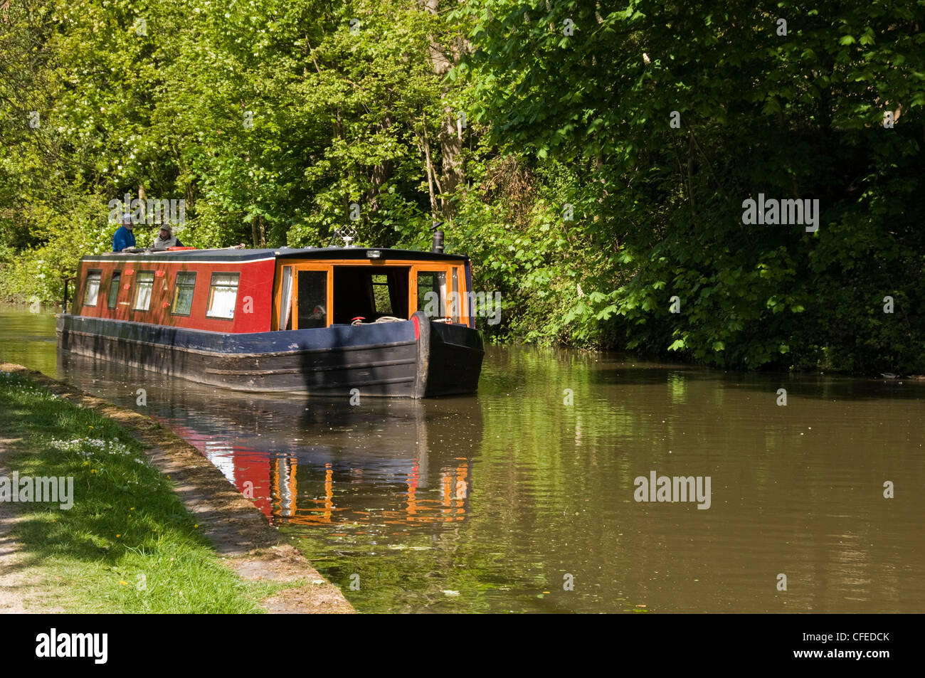 Narrowboat travelling along sunny scenic rural stretch of water (Leeds Liverpool Canal) with couple on board - Bingley, West Yorkshire, England, UK Stock Photo