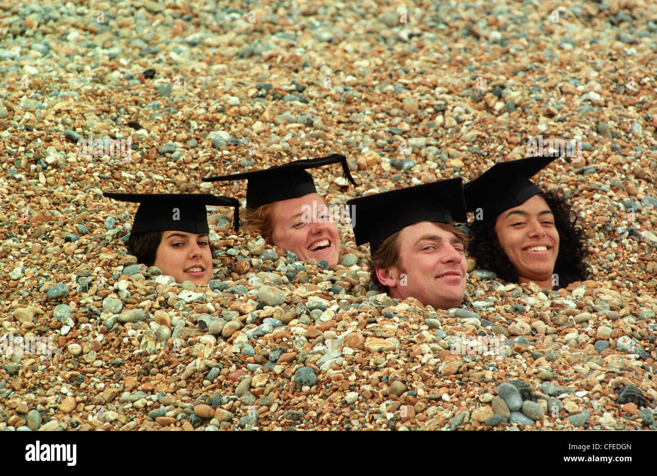 Three female graduates wearing mortar boards celebrate their degrees from the University of Brighton with a happy fellow male graduate buried up to their necks in the shingle on Brighton beach. Stock Photo