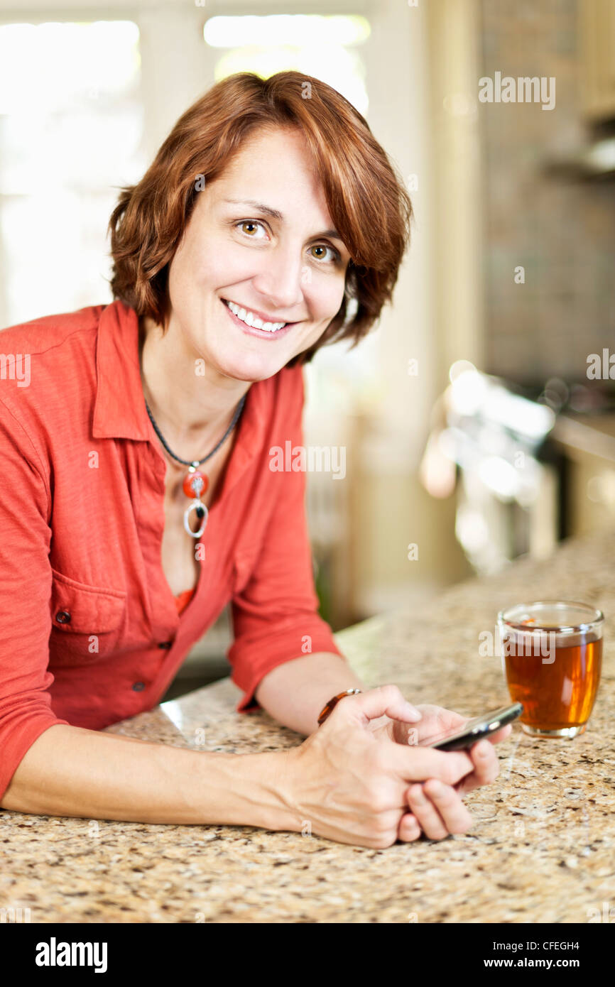 Smiling mature woman texting on phone in kitchen at home Stock Photo