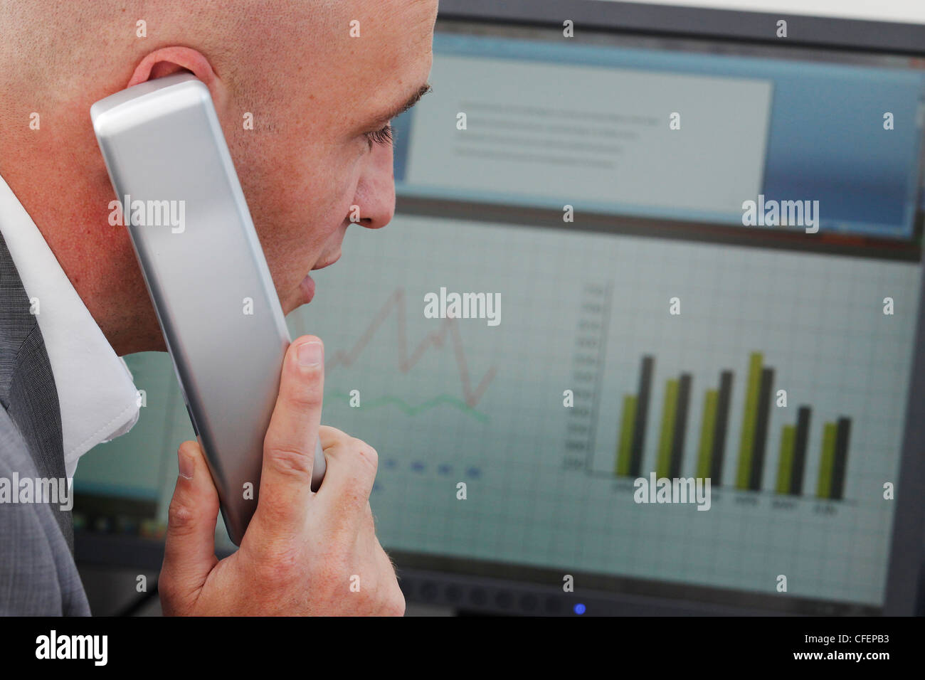 Model poses as an office worker speaking on the phone in front of a computer monitor showing graphs and charts Stock Photo