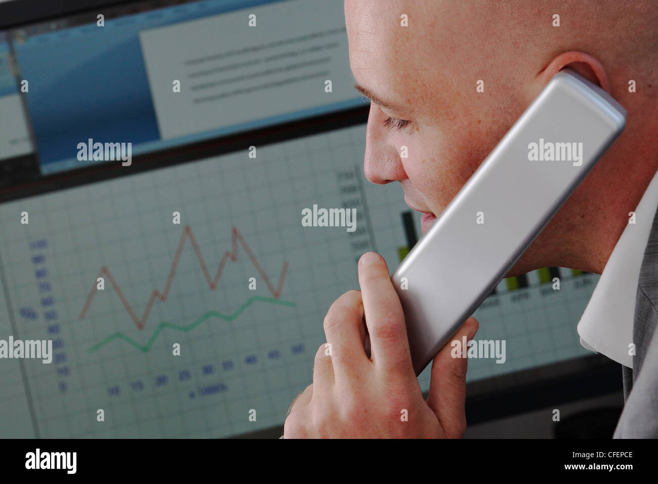 Model poses as an office worker speaking on the phone in front of a computer monitor showing graphs and charts Stock Photo