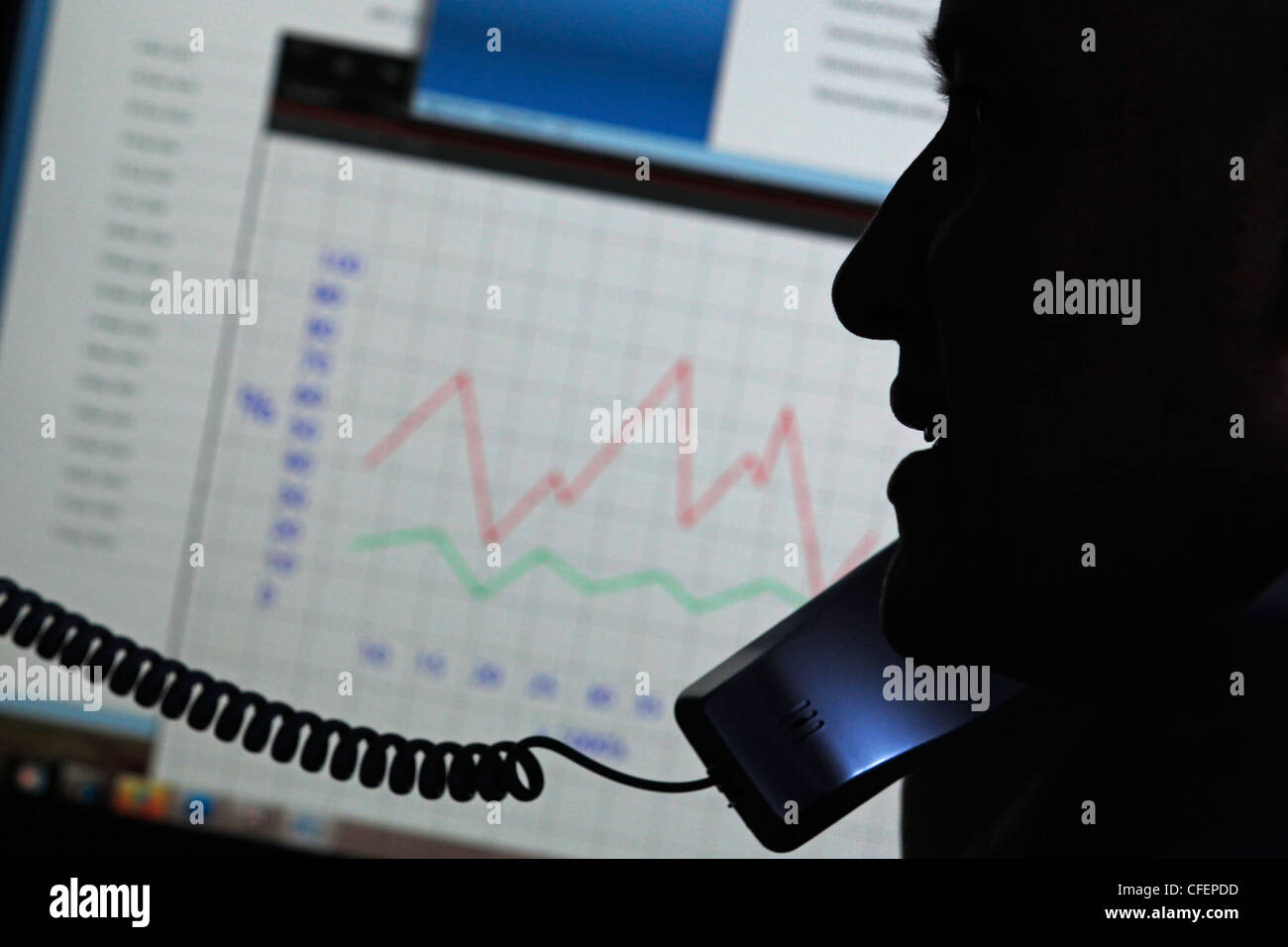 Model poses as an office worker speaking on the phone in front of a computer monitor showing graphs and charts Stock Photo