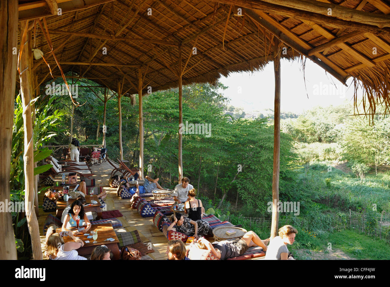 Travellers relaxing at reataurant terrace under palm roof, Nam Khan river, tributary to Mekong river, Luang Prabang, Laos Stock Photo