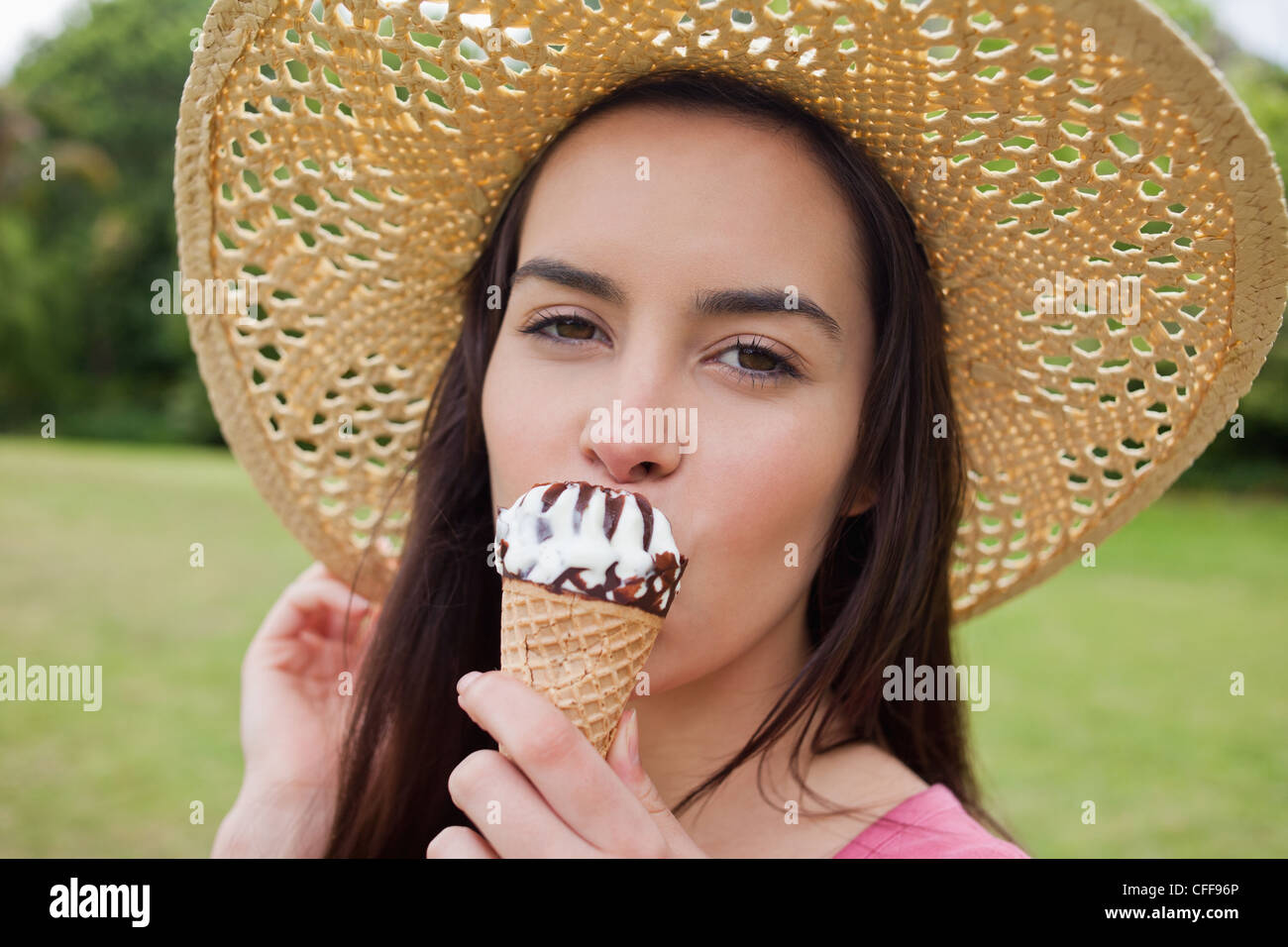 Young woman standing in a park while wearing a hat and eating an ice