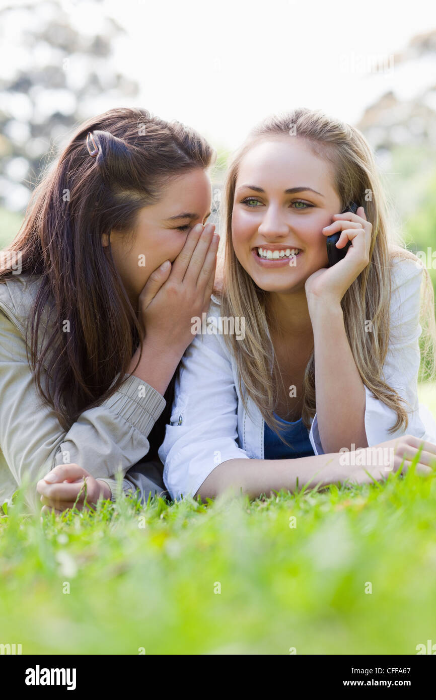 Young smiling woman calling while lying on the grass and being told a secret Stock Photo