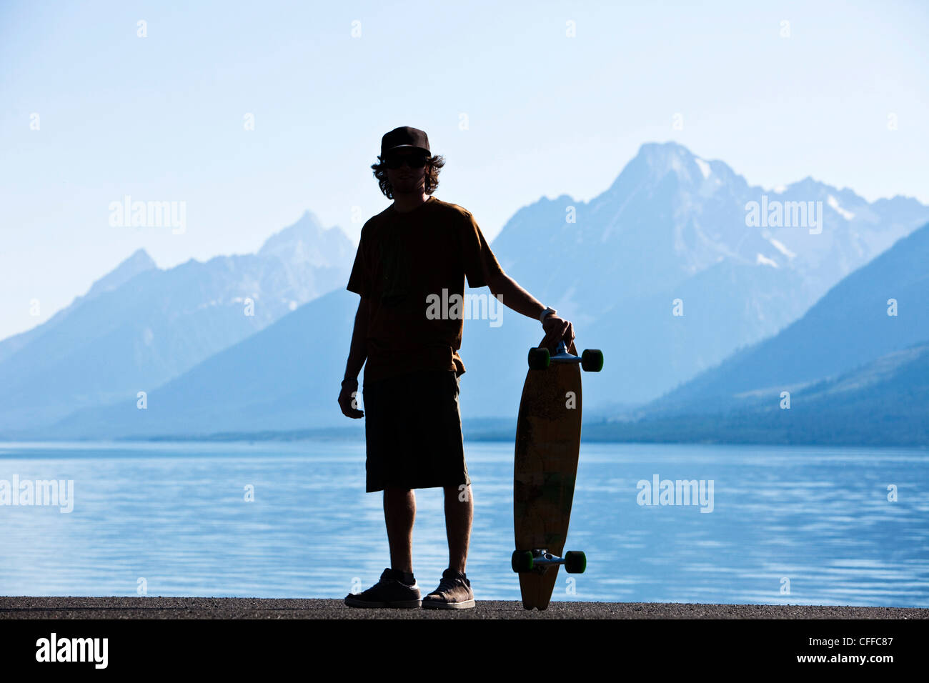 A young man standing next to his long board next to a lake in Wyoming. Stock Photo