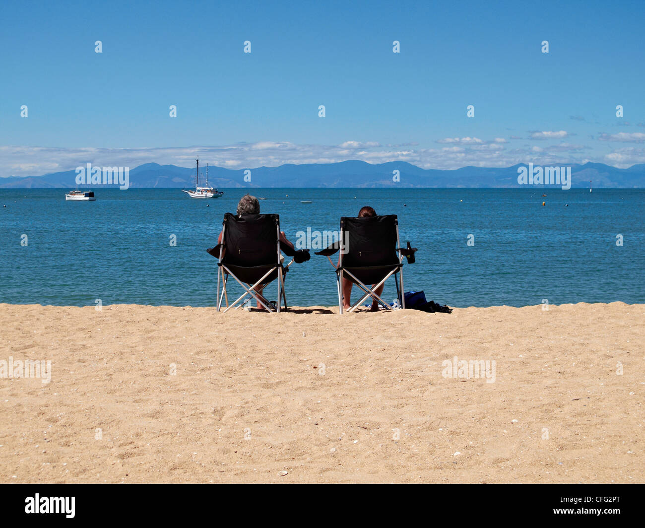 2 two people relax sunbathing on Kaiteriteri beach in New Zealand looking out towards the sea to the mountains Stock Photo
