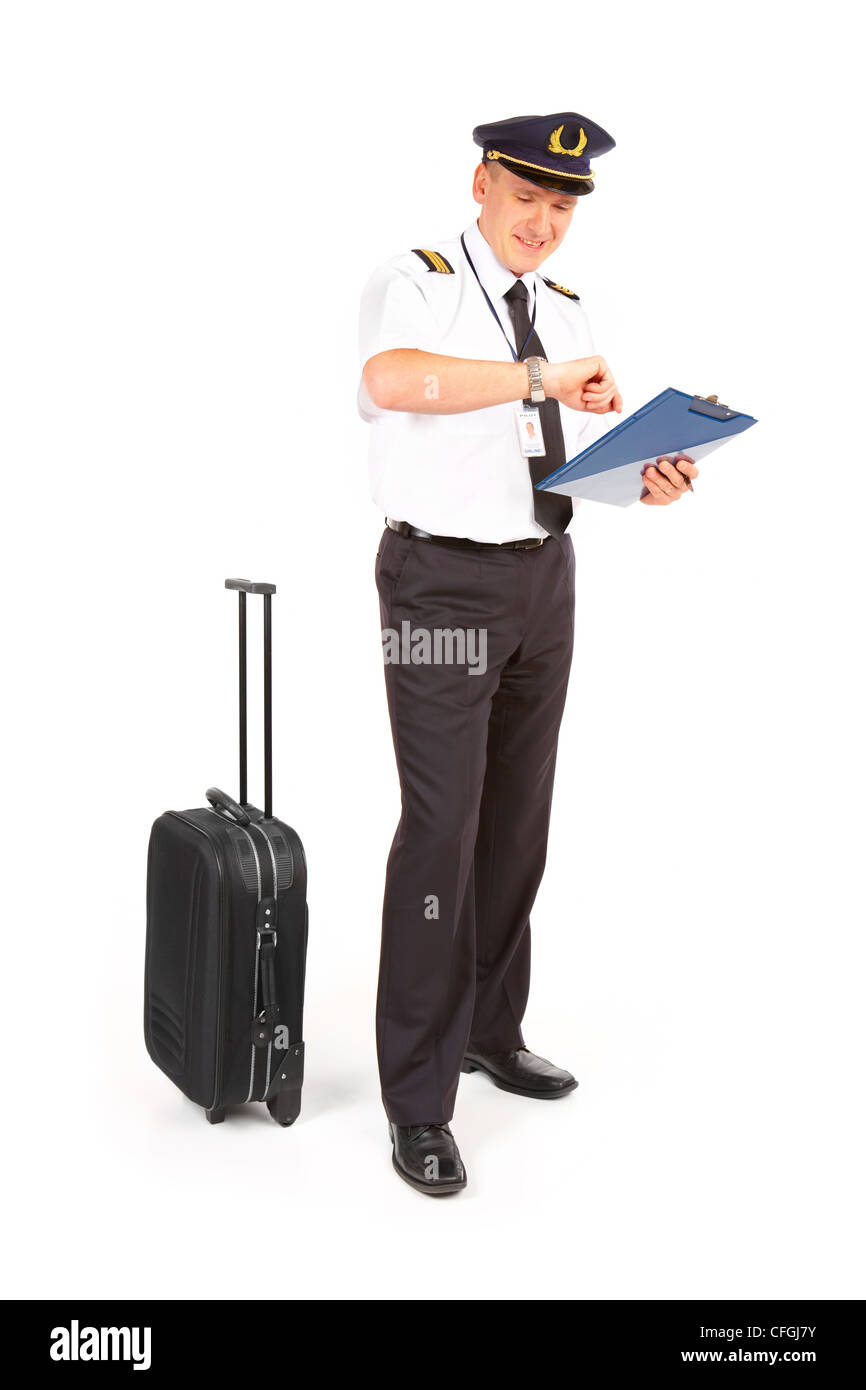 Cheerful pilot wearing uniform with epaulets standing with trolley bag and documents, checking time isolated on white background Stock Photo