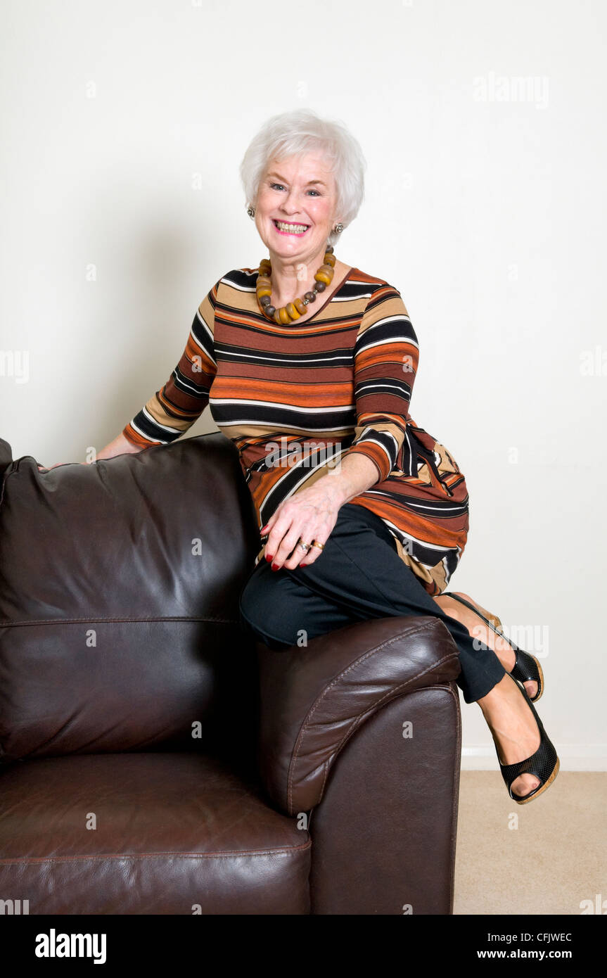 Attractive senior lady sat on edge of leather sofa smiling and looking relaxed Stock Photo